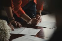 A person signing paperwork with a pen at a desk while another person points to the document